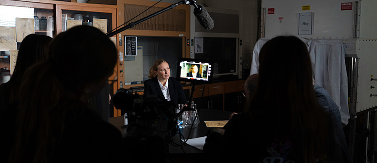 Sillhouettes work a camera and sound boom while filming a young woman in a science lab