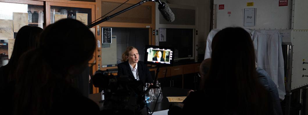 Woman being filmed during an interview in a lab setting, with a camera crew and lighting equipment visible.