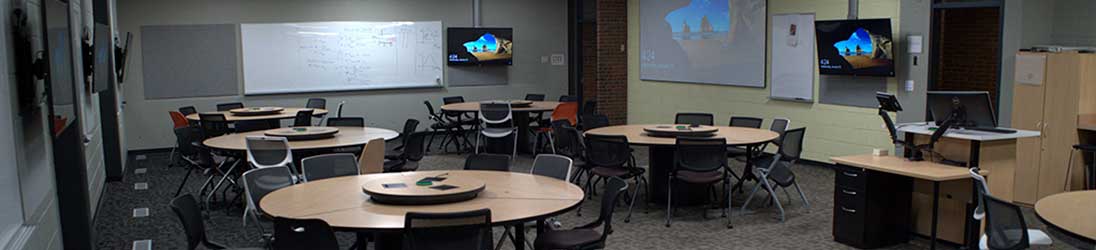Classroom with instructor podium, group tables, video displays on the walls, and a whiteboard