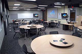 Classroom with group tables, a projector, video displays on the walls, and white boards
