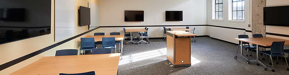 Classroom with five tables for groups, five video display screens on the walls, and a center instructor podium