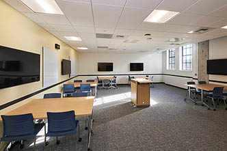 Classroom with five tables for groups, five video display screens on the walls, and a center instructor podium